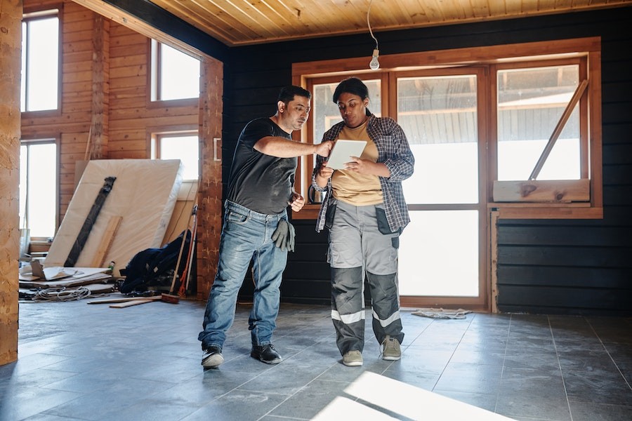 Man and woman standing in a house that’s being renovated. He points to a tablet she is holding.